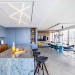 Modern open-concept kitchen and dining area with a marble waterfall island, sleek blue cabinetry, floor-to-ceiling windows, contemporary lighting, and panoramic city views.