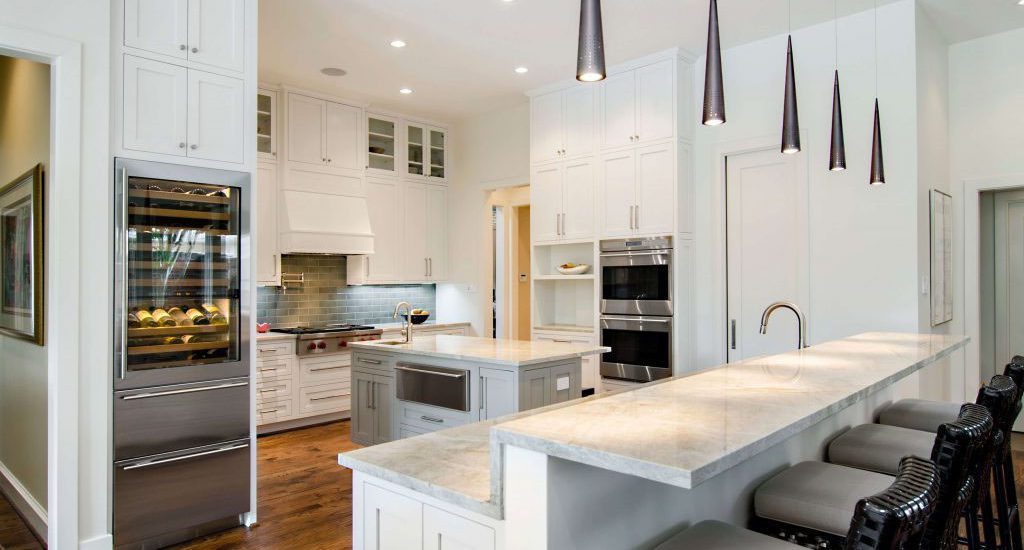 Modern, spacious white shaker kitchen remodel featuring a large breakfast bar with barstools, stainless steel appliances, a built-in wine cooler, hardwood floors, and contemporary pendant lighting.