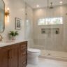 Bright modern bathroom addition with wood vanity, brass fixtures, glass shower, light tile, and natural daylight.