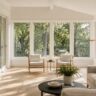 Sunlit home addition with large windows, pale oak floors, neutral seating, and brass floor lamp overlooking trees.